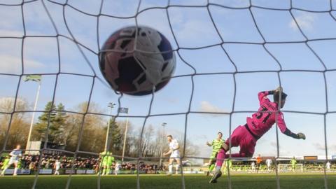 A goalkeeper attempts to make a save but the ball is already beyond him and in the net