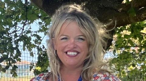 A headshot of a smiling blonde woman in a floral dress. There is a tree and a wire fence in the background