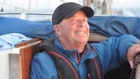 A man in a blue jacket, black vest and black hat. He is sitting in a boat.