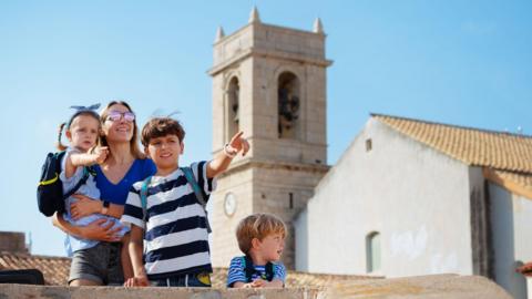 A woman wearing a tshirt, shorts and sunglasses holds a young girl wearing a backpack while two young boys stand in front of her looking and pointing into the distance. They're in front of a white bell tower in a European town.
