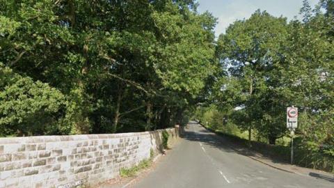 The entrance to Dawson Lane, with a sign on the left along with a stone wall and trees lining the opposite side of the road. There is a sign saying 'except for access' on the right hand side, with an image of a lorry and a 7.5 tonne vehicle limit on it.