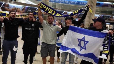 Maccabi Tel Aviv fans hold their team's flag and the Israeli flag and celebrate in a stadium.