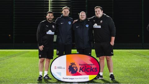 Four men, two wearing Southampton football club tracksuit kits (from the Saints Foundation) and two wearing Portsmouth Football club tracksuit kits (From Pompey in the Community). They are stood in a row with their arms around each other on a green astro turf pitch with a sign in front of them which reads "Premier League Kicks" 
