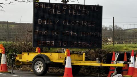 A large matrix sign on a yellow trailer giving details of the road closures. There are traffic cones around it and green fields and hills in the background.