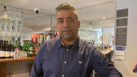 Restaurant owner Ashraf Ali is standing behind the bar. He has dark hair in a small quiff. He is wearing a blue shirt with a small white motif on it. There are bottles and glasses on the mirrored bar behind him. 