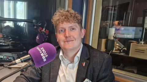 A young man with blond curly hair wearing a grey suit sits in a radio studio and smiles at the camera. He is sat beside a purple microphone that reads "BBC Radio Stoke" on the side in white writing.