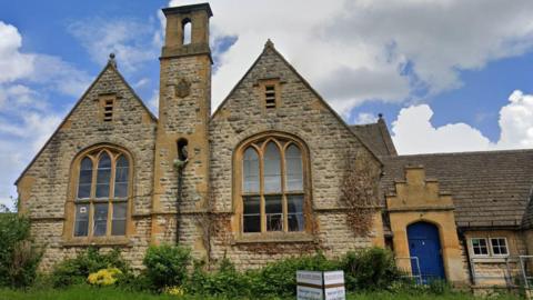 An old schoolhouse, built in Cotswold stone, with two pointed gables, large windows and a central chimney-like pillar between the two. There is a door painted blue besides one of the gables. There is a green space with grass and bushes in front of the building, and a sign. It is a partly cloudy day, with sections of blue sky and cloud.