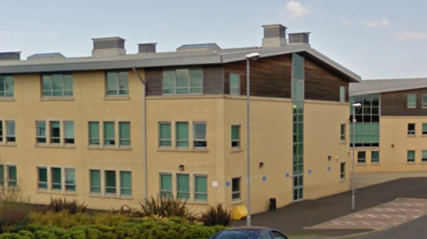 Beath High School is a modern three-storey yellow building with a brown top section. It has greenery off to the left and a pavement and road with streetlights on one side.