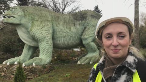 Lydia Lee standing in front of one of the dinosaur sculptures, wearing a hardhat and hi-vis jacket