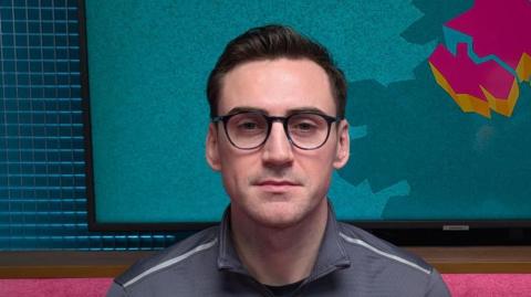 A man is looking straight at the camera. He has short jet black hair, which is slicked back. He has black-rimmed glasses. He is wearing a grey sports top with a zip. He is sitting in a podcast studio with a pink sofa, behind his head is a TV with the logo for The State of Us podcast, which is a teal background with a stylised image of Northern Ireland in pink.