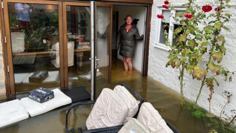 Nicky Vernede stands at the doorway to her patio. There is water above her ankles which is inside and outside the house.