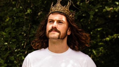 Ward is seen long curly hair and sideburns wearing a gold crown and a white T-shirt looks upward while standing outdoors, with green leaves and branches in the background.