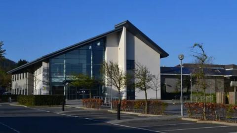 The Sixth Form Centre is a modern architectural building with a slanted roof and large glass windows dominating the front facade. The area around the building has paved walkways, parking spaces and landscaped greenery.