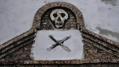 An artistic impression of a skull appears in some brickwork above a black 'X' on greay plaster.