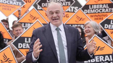 Liberal Democrat leader Sir Ed Davey stands in front of a group of supporters in Guildford, Surrey, holding orange Liberal Democrat signs.