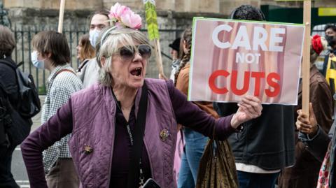 A lady with grey hair and a headband with pink roses on top of it holds a sign which reads: "Care Not Cuts". She is wearing a purple jumper and a purple quilted waistcoat. Other protestors are visible in the background. 