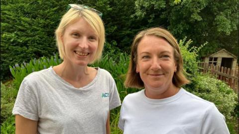 Two women face the camera and smile. Behind them is a variety of green foliage and a small shed. The woman on the left wears a grey t-shirt and has glasses on her head. The woman on the right wears a white t-shirt