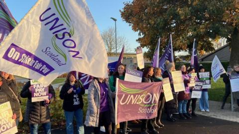 A group of men and women holding banners stand on a picket line outside a school.