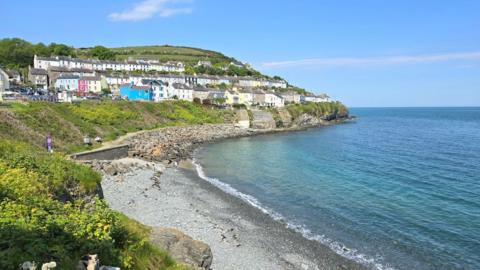 view from cove of shingle and sea, hilly coastline with white and colourful houses on hill in background, surrounded by trees