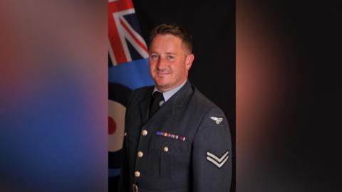 RAF veteran David Morris is dressed in a navy blue military uniform. He smiles as he looks at the camera. A Union Jack flag can be seen in the background of the photo. 