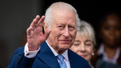 King Charles, dressed in a blue coat, white shirt and blue tie, holds up his right hand to wave, during his 76th birthday after the opening of the first Coronation Food Hub in Deptford. 