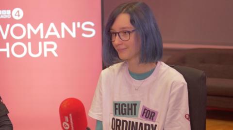 Katie Nellist sits in the Woman's Hour studio, wearing glasses a white t-shirt that says Fight For Ordinary