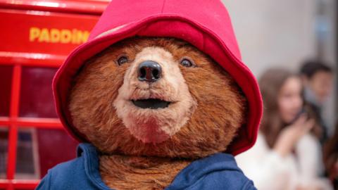 Paddington Bear wearing a blue duffle coat and a red hat in front of a British red phonebox