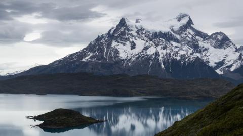 A cold and snowy mountain. Still water is at the foot of the mountain. It is a cloudy day.