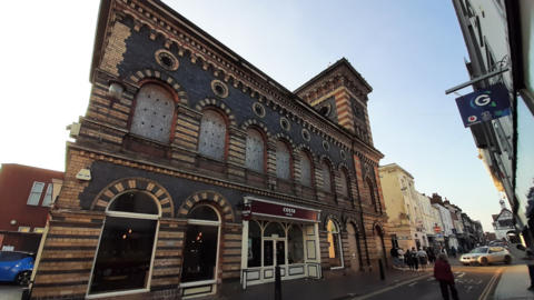 The orange and black building stands prominently on a street and a Costa coffee house can be seen inside the building. The upper level is boarded up and a person is walking down the street alongside the building.