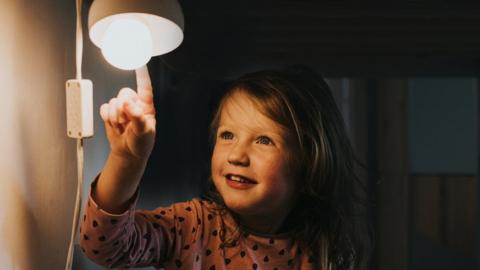 young girl in pyjamas, in the dark, smiling and pointing at a little night light. Darkness provides a space for copy. Conceptual image.