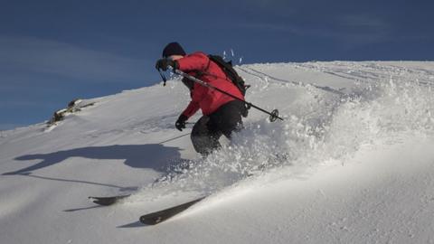 A man in a red jacket skiing on white snow against a blue sky, snow sprays from his skis