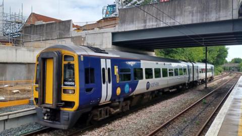 A blue, white and yellow Northern train passing through a station. It is made up of two carriages and there is a concrete bridge above it and building work behind it.