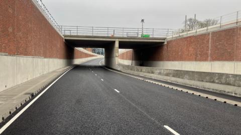 Image of the newly built underpass. There are walls on either side. The left hand side of the road surface is visible in the shot with freshly painted white lines running along it.