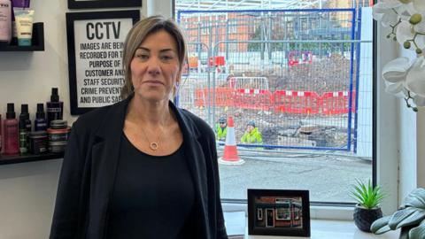 Catherine Prior had long brown hair and is wearing a black top and a black jacket. She is standing in front of her hair salon window through which roadworks can be ween with two workers in high visibility yellow jackets and hard hats.