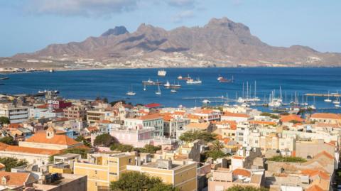 View over Mindelo, Sao Vicente, Cape Verde showing buildings with terracotta-coloured roofs in front of the blue ocean, which has several boats in it. Behind the ocean is a grey mountain and the sky is light and blue.