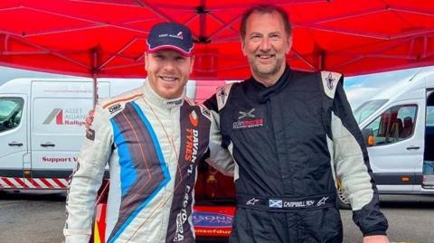 Sir Chris Hoy and Campbell Roy stand arm in arm, wearing drivers outfits with two vans in the background. They are standing under a canopy.