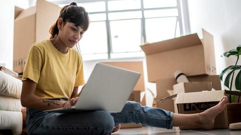 Person inside using laptop while sitting on floor with cardboard boxes in living room after moving house