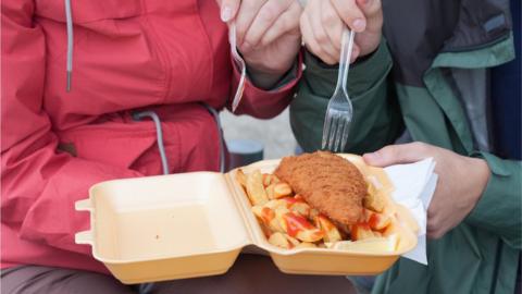 Couple eating fish and chips using a plastic fork from a single-use tray.