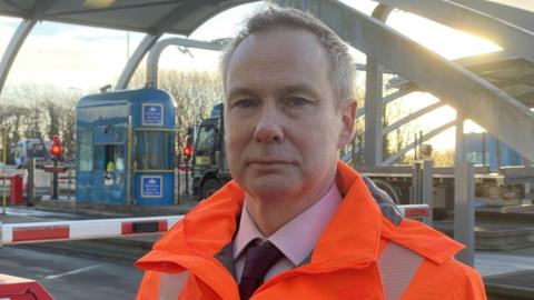 A man with short fair hair looks into the camera as he stands in front of toll booths at the entrance to a bridge. He is wearing a hi-vis orange jacked over a suit, lilac shirt and dark tie. Behind him is a red and white barrier blocking a road. A blue toll booth and red lights can also be seen. A truck is waiting at the toll booth. The sun is low in the sky.