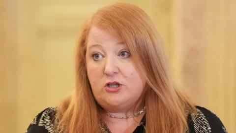 Naomi Long is wearing a black and white t shirt and a necklace. She has ginger hair. The background is blurry. 