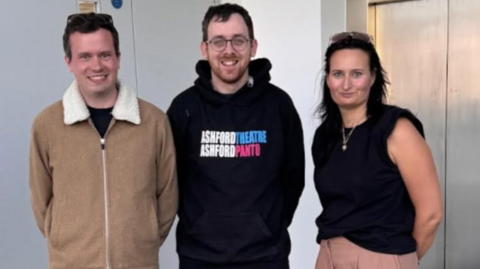 Two men standing next to a woman. One man is wearing a brown jacket and the other is wearing a black hoodie which says "Ashford Theatre. Ashford Panto" on it. The woman is wearing a black shirt with no sleeves.