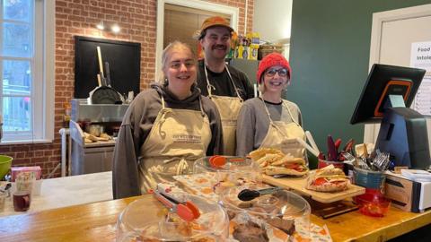 Volunteers Ellie and Jock standing next to Jo Gordon behind the till at the Willow Tree Coffee and Kitchen. Pastries and sandwiches are displayed on the counter in front of them. They are all wearing white aprons. Ellie has straight blonde hair tied in a ponytail. Jack is wearing a black cap and has moustache. Jo has short, straight grey hair and she is wearing round glasses and a red beanie.