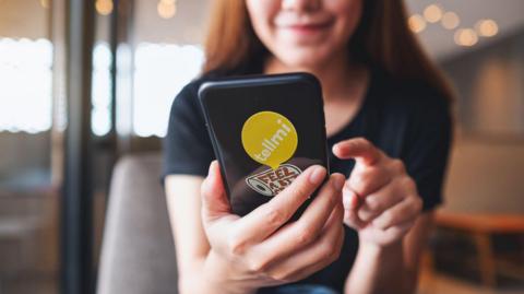 A teenage girl seen to use a phone with a TellMe sticker at the back. She is smiling. She seems to be sitting in a public space.