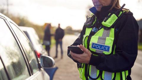 A woman police officer deals with a traffic incident and uses a mobile phone. Her uniform and the ends of her hair are visible. A car is parked on the verge and people are standing nearby.