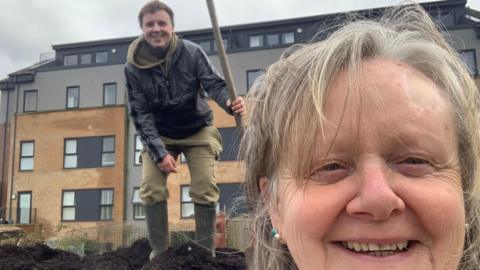 Sara, front right, taking a selfie photo, while Noah, rear, stands and smiles. He is holding a spade, and the pair are standing in front of an apartment block. Sarah is older and has grey hair, Noah has a black jacket, green trousers and wellies on