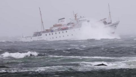 A white vessel struggles through big waves, her bow is up in the air as she hits a large wave, a plume of spray billows through the air.