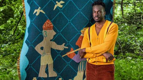 Cameroonian-Nigerian interior designer Amechi Mandi, dressed in a yellow cardigan and burnt brown cords, stands in a garden with arms folded in front of one of his throw blankets called 'Chief Palaver' showing two figures and two birds stitched on to blue diamond designed thick woven fabric 