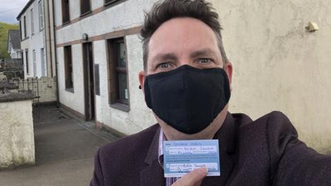BBC Wales' George Herd holds up vaccine record outside his GP surgery in Gwynedd