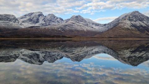 Snow dusted mountains and clouds in a blue sky reflected in a loch