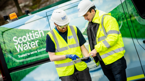 Two Scottish Water workers in high-visibility jackets and hard hats stand next to a Scottish Water van